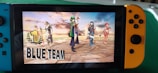 A Nintendo Switch console displays a victory screen from a video game, featuring animated characters standing triumphantly on a stone platform with a dramatic sky in the background. The words '1 Blue Team' are prominently displayed on the screen.