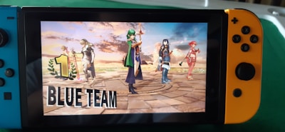 A Nintendo Switch console displays a victory screen from a video game, featuring animated characters standing triumphantly on a stone platform with a dramatic sky in the background. The words '1 Blue Team' are prominently displayed on the screen.