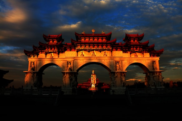 A traditional Chinese gate with ornate decorations is illuminated against a dramatic sky filled with clouds. The large arched structure features intricate carvings and includes a prominent golden statue in the center.
