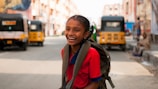 A young girl wearing a red school uniform and a backpack is smiling brightly while standing on a street. In the background, there are several auto-rickshaws parked on either side of the street. Buildings line both sides of the road, and the scene conveys a sense of a busy, urban environment.