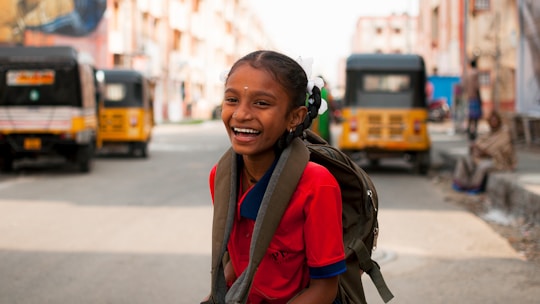 A young girl wearing a red school uniform and a backpack is smiling brightly while standing on a street. In the background, there are several auto-rickshaws parked on either side of the street. Buildings line both sides of the road, and the scene conveys a sense of a busy, urban environment.