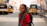 A young girl wearing a red school uniform and a backpack is smiling brightly while standing on a street. In the background, there are several auto-rickshaws parked on either side of the street. Buildings line both sides of the road, and the scene conveys a sense of a busy, urban environment.