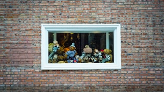 A variety of stuffed animals are arranged in a display inside a window with a white frame, set against a brick wall. The collection includes bears, dogs, and other plush toys.