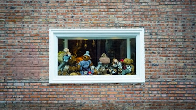 A variety of stuffed animals are arranged in a display inside a window with a white frame, set against a brick wall. The collection includes bears, dogs, and other plush toys.