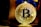 A golden Bitcoin coin with intricate circuit-like details is prominently displayed. It is held up by a figure wearing a blue costume.