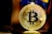 A golden Bitcoin coin with intricate circuit-like details is prominently displayed. It is held up by a figure wearing a blue costume.
