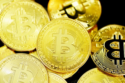Several gold coins with the Bitcoin symbol prominently displayed across their surfaces. The coins are layered and overlapping, creating a rich and textured appearance.