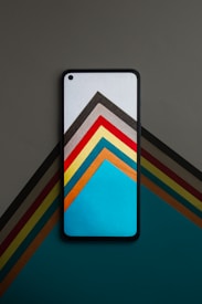 A smartphone is placed on a flat surface, displaying a colorful geometric pattern that appears to extend seamlessly onto the surface beneath it. The pattern consists of triangles and stripes in various colors, including blue, red, yellow, and brown.