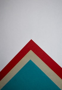 A geometric composition featuring three overlapping triangles arranged in descending order on a plain background. The triangles are colored red, beige, and teal and are perfectly aligned at their vertices.