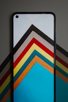 A smartphone with a screen displaying a series of colorful, geometric shapes layered in an abstract fashion. The background also shows similar geometric patterns that complement the phone's display. The shapes consist of a variety of colors arranged in a symmetrical pattern, forming visually striking arrows or chevrons.