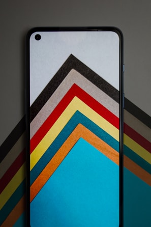 A smartphone with a screen displaying a series of colorful, geometric shapes layered in an abstract fashion. The background also shows similar geometric patterns that complement the phone's display. The shapes consist of a variety of colors arranged in a symmetrical pattern, forming visually striking arrows or chevrons.