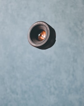 A single camera lens appears to float against a textured blue background. The lens is positioned in the upper left quadrant of the image, with its reflective glass surface visible.