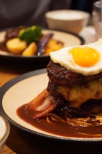 A gourmet dish featuring a layered stack of food, with a cooked egg on top displaying a bright yellow yolk. Beneath the egg, there is a slice of melted cheese, layers of meat, and bacon, all covered in a rich brown sauce. The dish is served on a round plate with another plate visible in the background containing potatoes and greens.