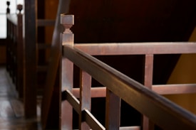 A close-up view of a wooden staircase railing with a geometric design. The image has a warm tone due to the natural lighting, and the focus is on the details and texture of the wood. The background is slightly blurred, allowing the railing to be the main subject.