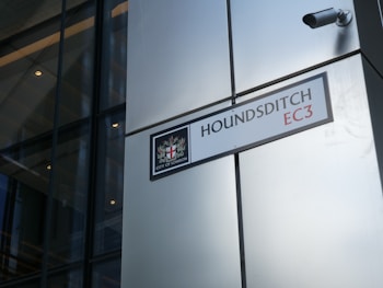 A street sign labeled 'HOUNDSDITCH EC3' is mounted on a modern building with reflective glass panels. The sign includes the City of London emblem. A security camera is positioned above the sign.