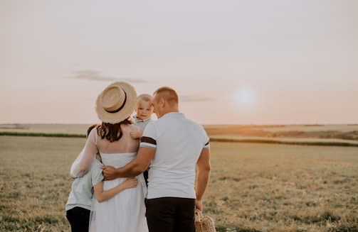 A family of four stands together in a field watching the sunset. The adults, one wearing a straw hat, hold their children close. The scene is peaceful and pastoral, with a soft glow from the setting sun.