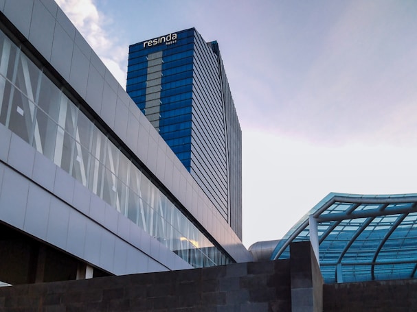 A modern, high-rise building with blue-tinted glass windows. The building has the name 'Resinda Hotel' on the top. To the right, there's a structure with a glass roof and metal beams. The sky is clear with a light blue hue, with a hint of clouds visible.