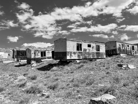 A series of dilapidated, abandoned trailers sit on a grassy hillside under a sky filled with scattered clouds. The trailers are weathered and rusted, with broken windows and a sense of decay. In the background, distant mountains are visible, adding to the remote and isolated atmosphere.