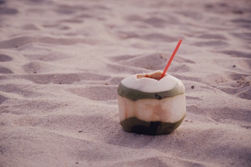 A coconut with a green outer shell rests on sandy beach terrain. A red straw is inserted into the top, suggesting it is being used as a drink container. The sand is light tan and appears soft, with gentle undulations.