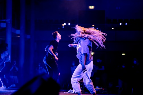 Two people are energetically dancing on a stage under vibrant blue and purple lighting. One dancer, with long, flowing hair, appears to be mid-movement, while the other is slightly turned and animated. The background shows a darkened audience and a hint of a building structure, creating a lively and dynamic atmosphere.