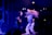Two people are energetically dancing on a stage under vibrant blue and purple lighting. One dancer, with long, flowing hair, appears to be mid-movement, while the other is slightly turned and animated. The background shows a darkened audience and a hint of a building structure, creating a lively and dynamic atmosphere.