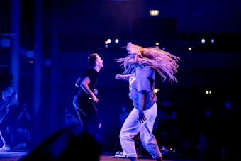 Two people are energetically dancing on a stage under vibrant blue and purple lighting. One dancer, with long, flowing hair, appears to be mid-movement, while the other is slightly turned and animated. The background shows a darkened audience and a hint of a building structure, creating a lively and dynamic atmosphere.