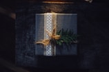 A neatly wrapped gift box featuring a textured dark cover with a polka-dotted ribbon tied around it. The package is adorned with a sprig of evergreen, giving it a rustic and natural appearance. The background is a dark wooden surface, complementing the earthy and cozy aesthetic.