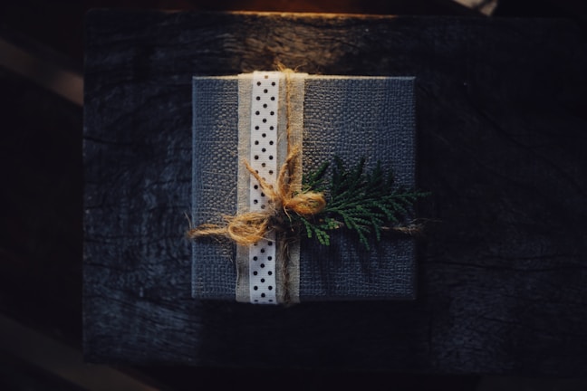 A neatly wrapped gift box featuring a textured dark cover with a polka-dotted ribbon tied around it. The package is adorned with a sprig of evergreen, giving it a rustic and natural appearance. The background is a dark wooden surface, complementing the earthy and cozy aesthetic.
