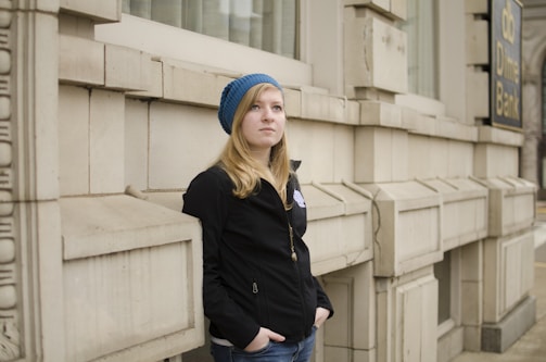 A young person with long hair stands against the corner of a stone building, wearing a blue knitted beanie and a black jacket. The setting appears urban, with part of a 'db Dime Bank' sign visible in the background.