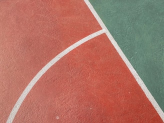 A section of a sports court with a rough textured surface. The court is divided into two parts: red and green, separated by white curved lines. The red section covers the majority of the image while the green section occupies the upper right corner.