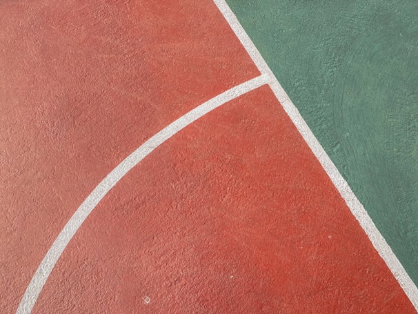 A section of a sports court with a rough textured surface. The court is divided into two parts: red and green, separated by white curved lines. The red section covers the majority of the image while the green section occupies the upper right corner.