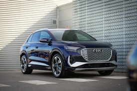 A sleek, modern SUV with a glossy blue finish is parked in an industrial setting with metal siding. The vehicle features a prominent front grille and aerodynamic design, reflecting the surrounding environment. The scene suggests a combination of style and functionality.