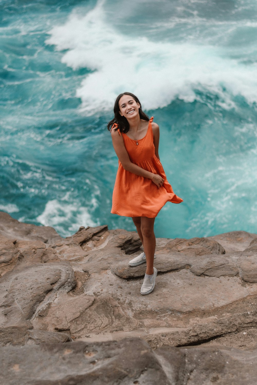 woman in orange sleeveless dress standing on rock formation near body of water during daytime