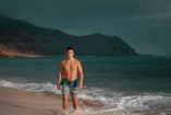 topless man in blue denim shorts standing on seashore during daytime
