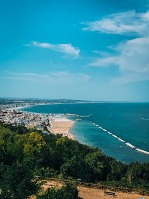 A panoramic view of a coastal city bordered by the sea, with a long stretch of sandy beaches lined with sun loungers and umbrellas. The water is a vivid blue, contrasting with the green trees in the foreground. White clouds are scattered across a bright blue sky.