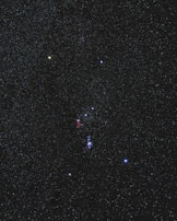 A clear night sky filled with numerous stars, dominated by a prominent constellation. The stars vary in color and brightness, with some appearing more luminescent. A faint reddish nebula is visible near the center of the image.