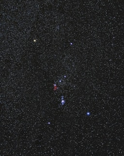 A clear night sky filled with numerous stars, dominated by a prominent constellation. The stars vary in color and brightness, with some appearing more luminescent. A faint reddish nebula is visible near the center of the image.