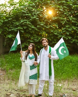 A couple dressed in white and green stands outdoors, each holding a flag with a crescent and star, symbolizing Pakistan. They are surrounded by lush greenery with sunlight peeking through the trees, creating a serene backdrop.