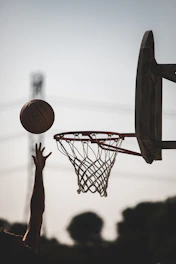 A person reaching toward a basketball flying toward a hoop against a bright sky