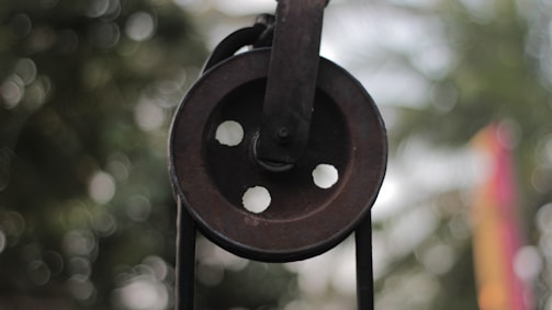 A close-up of an old, rusty pulley wheel with four circular holes. The background is blurred with a bokeh effect, featuring hints of green and other colors.