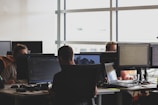 Several people working at desks with computer monitors, focusing on programming and other tasks. The office space is bright with natural light from large windows.
