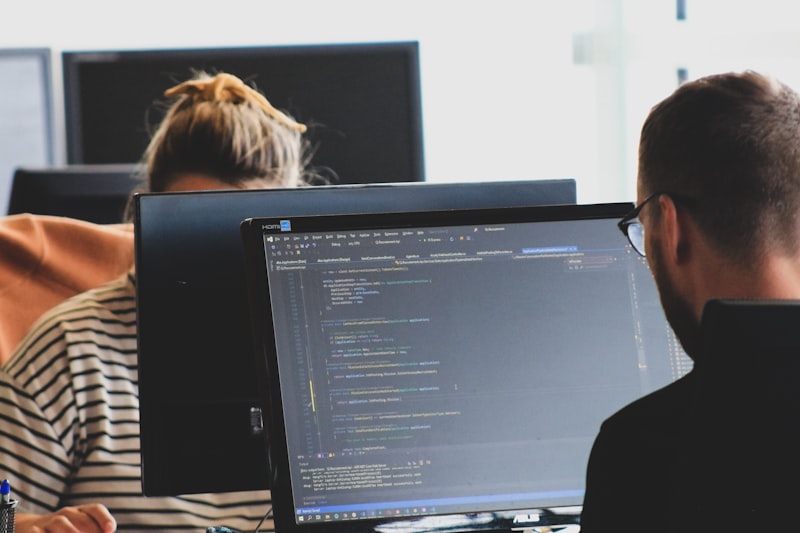 Two people working on computer code at monitors in a bright office workspace