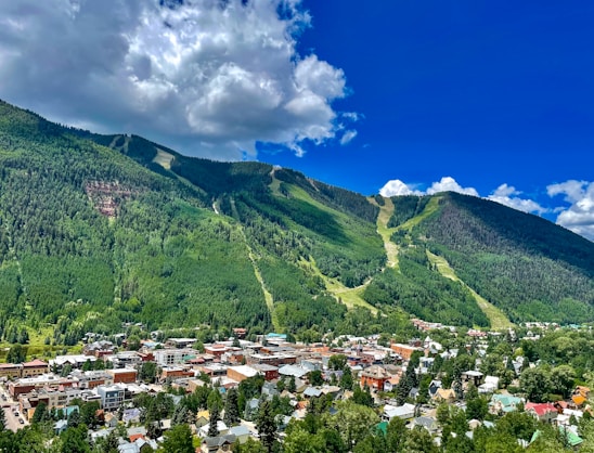 A vibrant town nestled at the base of lush green mountains, under a partly cloudy blue sky. The town is filled with various buildings, houses, and tree-lined streets. The mountains in the background are covered with dense forest and cut-through trails, possibly for skiing or hiking.
