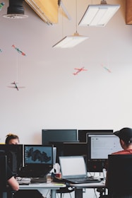 An office environment with multiple people working at desks equipped with computers. Hanging from the ceiling are small, model airplanes creating a whimsical touch. The lighting is provided by rectangular ceiling lights, and there are several computer monitors visible.