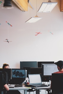 An office environment with multiple people working at desks equipped with computers. Hanging from the ceiling are small, model airplanes creating a whimsical touch. The lighting is provided by rectangular ceiling lights, and there are several computer monitors visible.