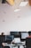An office environment with multiple people working at desks equipped with computers. Hanging from the ceiling are small, model airplanes creating a whimsical touch. The lighting is provided by rectangular ceiling lights, and there are several computer monitors visible.