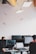 An office environment with multiple people working at desks equipped with computers. Hanging from the ceiling are small, model airplanes creating a whimsical touch. The lighting is provided by rectangular ceiling lights, and there are several computer monitors visible.