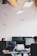 An office environment with multiple people working at desks equipped with computers. Hanging from the ceiling are small, model airplanes creating a whimsical touch. The lighting is provided by rectangular ceiling lights, and there are several computer monitors visible.