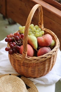 A wicker basket filled with fresh fruit, including green and red grapes, peaches, and a pear, placed on a white cloth. The basket is positioned next to a woven straw hat.