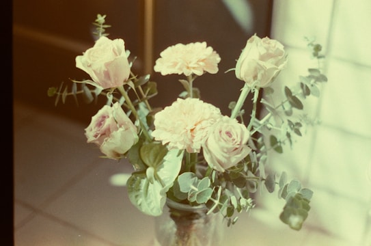 A bouquet of pastel-colored roses and carnations arranged in a glass vase. The flowers are surrounded by green eucalyptus leaves, and the setting includes soft lighting creating a dreamy and gentle atmosphere.